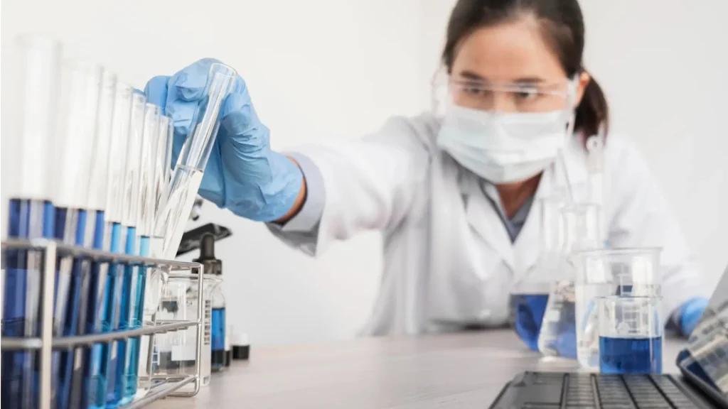 A scientist wearing a lab coat, mask, and safety goggles places a test tube into a rack filled with blue liquid samples.