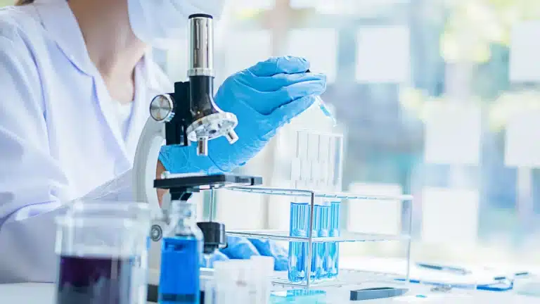 A scientist in a lab coat and blue gloves uses a pipette to add blue liquid into a test tube next to a microscope.