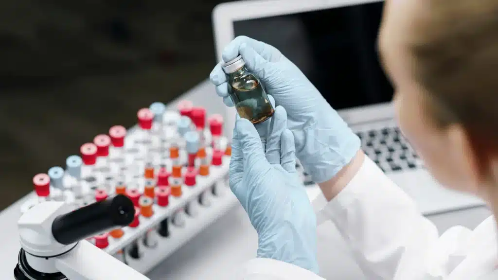 A scientist in blue gloves holding a glass vial, with a rack of test tubes and a microscope on the laboratory desk.