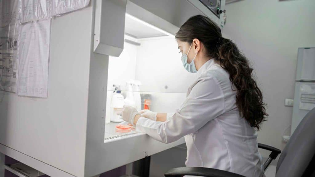 A scientist wearing a lab coat, mask, and gloves working inside a laboratory biosafety cabinet.