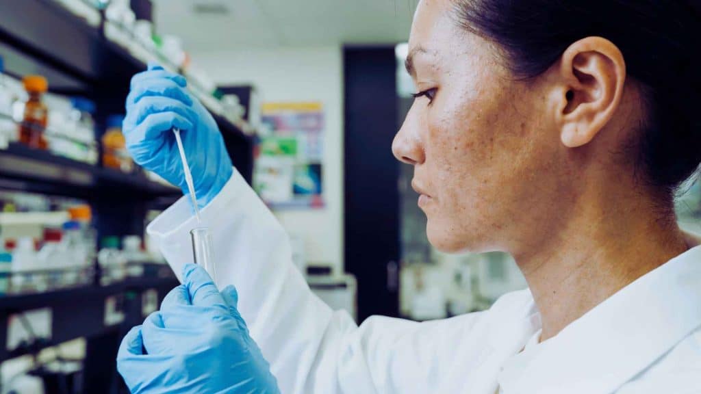 A scientist in a lab coat and blue gloves uses a pipette to transfer liquid into a test tube.