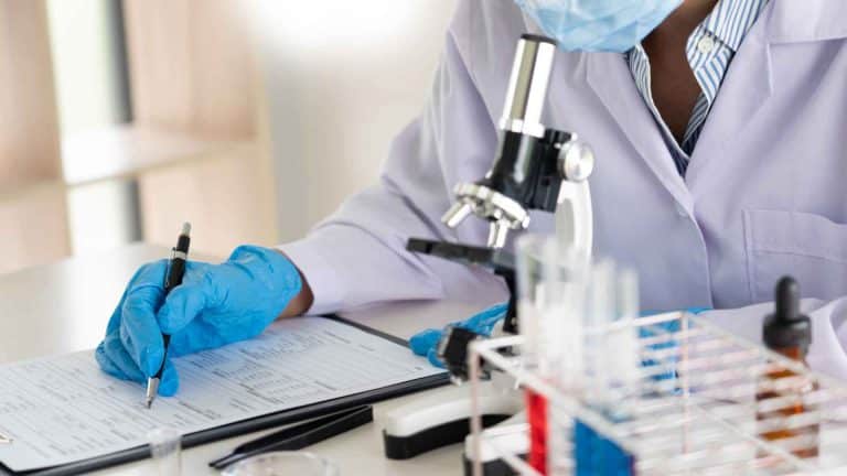 A scientist in a lab coat and blue gloves writes on a clipboard next to a microscope and a test tube rack.