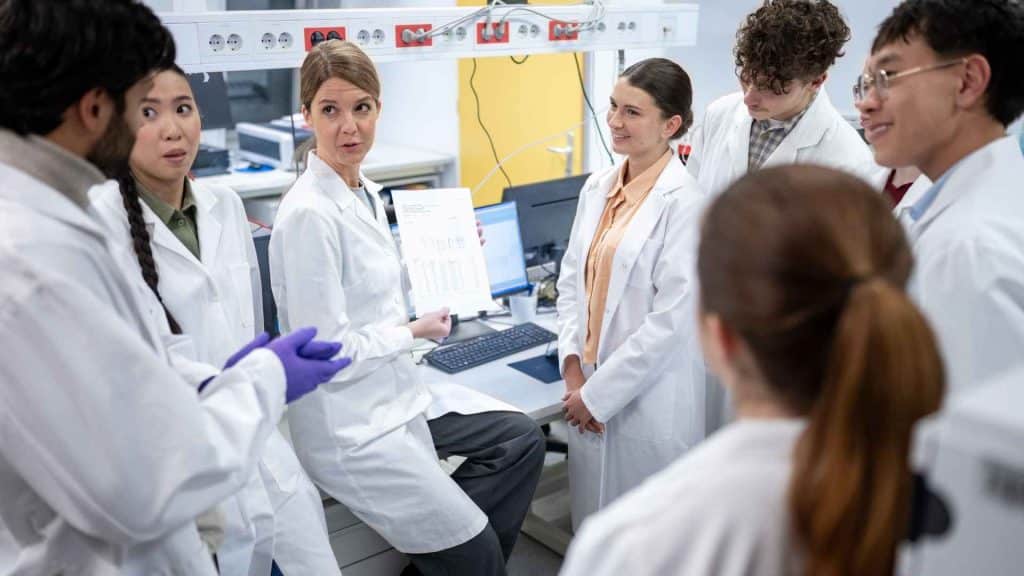 A group of scientists in lab coats gather around a colleague who is holding up a printed document for review in a laboratory.